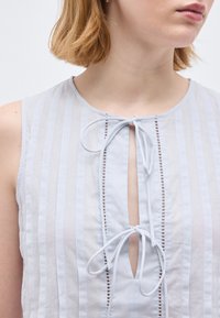 Γυναίκα που φοράει αμάνικο λευκό τοπ με ρίγες, δύο δεμένα φιόγκους στο κέντρο του ντεκολτέ και κάθετες λεπτομέρειες με ανοίγματα.