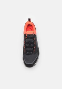 Scarpa sportiva nera con tomaia in mesh testurizzato, accenti arancioni brillanti, lacci e un manico. Presenta un collare imbottito e una suola in gomma.