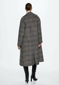 Long manteau à carreaux dans des tons de gris, présentant un design croisé et une longueur mi-mollet. Le matériau semble texturé avec une silhouette structurée.