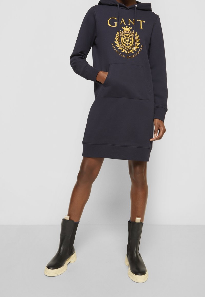 Personne portant une robe à capuche bleu marine avec un logo GANT jaune et des bottes noires aux semelles beiges épaisses sur un fond gris uni.