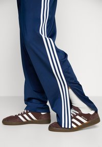 Pantalones deportivos de color navy con diseño de tres franjas blancas, que cuentan con tobillos con cremallera. Zapatillas marrones con franjas blancas y suelas texturizadas. Calcetines blancos visibles.