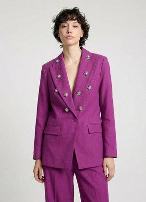 Donna che indossa una giacca doppiopetto viola con bottoni decorativi argento a forma di stella e pantaloni abbinati, in piedi davanti a uno sfondo semplice.