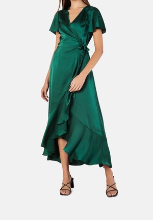 Kvinde iført en mørkegrøn satin slå-om-kjole med flagrende ærmer, asymmetrisk flæsekant og sorte sandaler med hæl og remme.