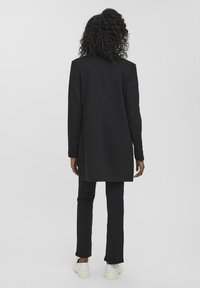 Blazer nero su misura con taglio dritto, maniche lunghe e apertura posteriore singola, abbinato a pantaloni neri a gamba dritta e sneaker bianche.