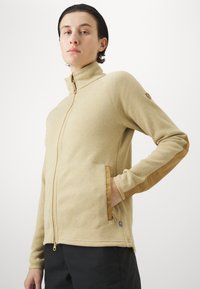 Giacca in pile beige con colletto alto, chiusura a zip e accenti in pelle sulle maniche. Presenta tasche laterali e un piccolo logo sulla spalla.