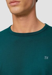 Türkiser Rundhalsausschnitt-T-Shirt aus weicher Baumwolle, mit einem kleinen Logo-Patch auf der linken Brust und einer glatten, gleichmäßigen Textur.