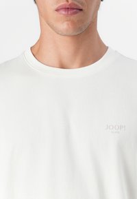 Hvit bomulls t-skjorte med rund hals, med en subtil lys grå "JOOP! JEANS" logo på venstre side. Glatt tekstur.