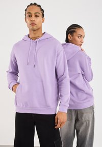 Due persone con capelli intrecciati che indossano felpe lavanda coordinate, una di spalle con le mani nelle tasche e l'altra girata.