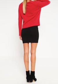 Czerwony, oversize sweter wykonany z miękkiego materiału, zestawiony z dopasowaną, czarną spódnicą i czarnymi botkami za kostkę. Sylwetka widoczna z tyłu.