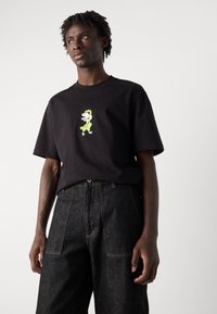 Camiseta de algodón negra con un gráfico de dibujos animados verde centrado en el pecho, combinada con pantalones de mezclilla oscuros. Manga corta, corte relajado.