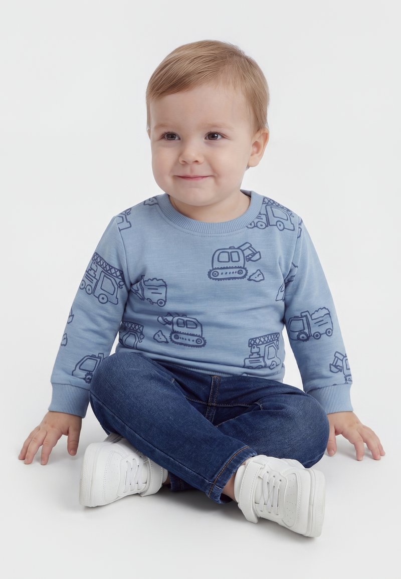 Tout-petit assis jambes croisées, portant un sweat-shirt bleu clair avec des motifs de véhicules de chantier, un jean foncé et des baskets blanches, souriant légèrement.