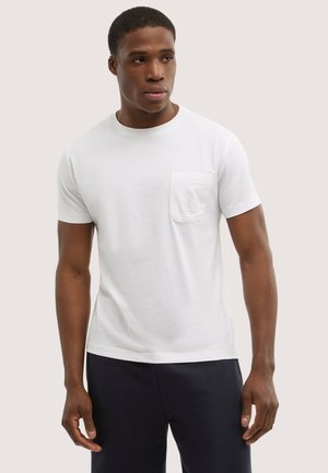 Uomo che indossa una semplice maglietta bianca a maniche corte con una piccola tasca sul petto sinistro e pantaloni scuri, che guarda di lato.