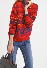 Červený vzorovaný svetr s horizontálními pruhy v oranžové a námořnické modři s žebrovaným lemem. Na spodní části je nápis „Moschino“ tučným modrým písmem.