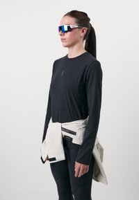 Black long-sleeve shirt with a logo, paired with black leggings. White utility jacket tied around the waist. Blue-tinted sport sunglasses.