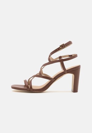 Sandales à talons hauts en cuir marron avec plusieurs brides croisées et une petite boucle. Le talon est de forme carrée et mesure environ 7,5 centimètres de haut.