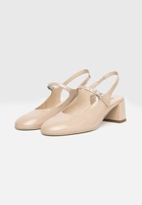 Zapatos de charol beige con talón abierto, punta redondeada, tacón bajo y correa ajustable en el tobillo con detalles de hardware metálico.