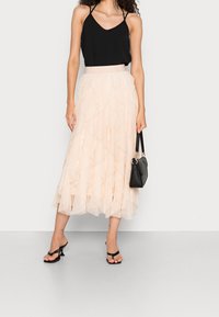 Haut noir sans manches associé à une jupe en tulle pêche à plusieurs couches, longueur genou. Le mannequin tient un sac à main noir et porte des sandales à talons noires.