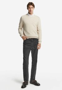 Jasno beżowy sweter dziergany z prążkowanymi mankietami, w zestawie z ciemnoszarymi dżinsami slim fit oraz ciemnymi butami z brązowymi sznurowadłami. Prosty design, bez wzorów.
