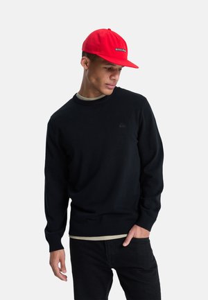 Young man wearing a red cap, black sweater, and black pants, standing with one hand in pocket, looking down to the side on a white background.