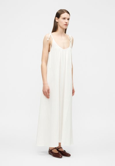 Γυναίκα ντυμένη με λευκό, αμάνικο μακρύ φόρεμα με δέσιμο στους ώμους και σκούρα καφέ παπούτσια τύπου Mary Jane, που στέκεται μπροστά σε απλό λευκό φόντο.