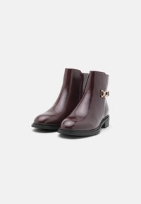 Bottes cheville en cuir bordeaux avec une finition lisse, fermeture éclair et détails en métal doré. Semelle plate noire. Design carré.