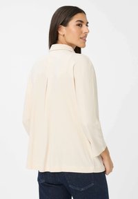 Femme aux cheveux bruns portant un blouse longue crème à manches longues et un jean foncé, se retournant sur son épaule sur un fond blanc.