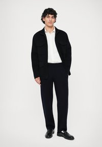 Young man standing wearing a black jacket, white shirt, dark trousers, and black shoes against a plain white background.