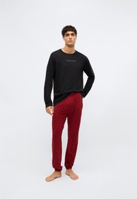 Melna garām piedurknēm krekls ar mazo Calvin Klein logotipu, apvienots ar sarkanām pidžamu biksēm ar smalku rakstu. Modelis stāv basām kājām.