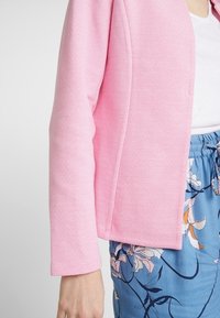 Žena v světle růžovém strukturovaném topu s dlouhým rukávem, který má kombinované modré květinové kalhoty se stahovací šňůrkou a bílou vnitřní košili.