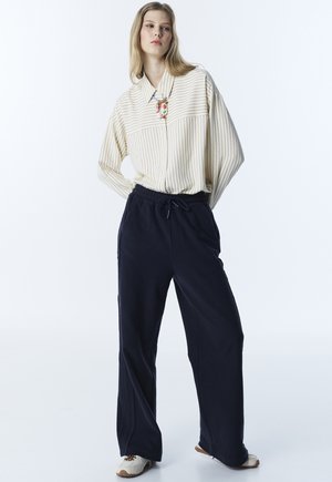 Femme blonde debout les mains dans le dos, portant une chemise beige rayée surdimensionnée, un pantalon large bleu marine, des baskets blanches et un collier coloré.