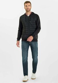 Svart hoodie med vadderad front, dragkedja och huva. Kombinerad med blå jeans och vita sneakers. Har en röd och vit krage. 