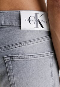 Los jeans de mezclilla gris presentan una etiqueta de piel blanca en la cintura con "Calvin Klein Jeans" en plata. Se pueden ver costuras sutiles y un bolsillo trasero.