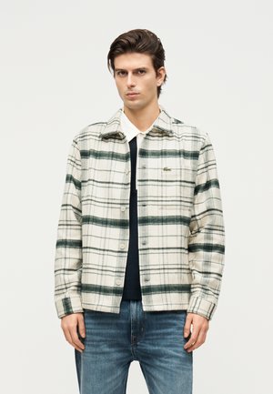 Mężczyzna ubrany w białą i ciemnozieloną kraciastą kurtkę zapinaną na guziki, na czarno-białą warstwową koszulę oraz niebieskie dżinsy, stoi na jednolitym tle.