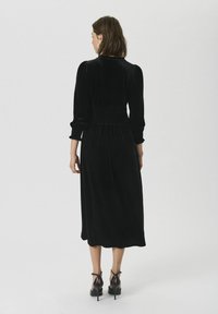 Schwarzes Samt-Midi-Kleid mit langen Ärmeln und gerafftem Bund. Das Kleid hat einen hohen Ausschnitt und eine fließende Silhouette. Dazu werden hochhackige Schuhe getragen.