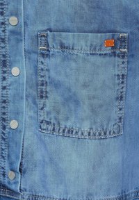 Světle modrá denimová košile se stříbrnými patentkami, jednou náprsní kapsou a malou oranžovou logovou štítkem. Textura vypadá hladce a umytě.