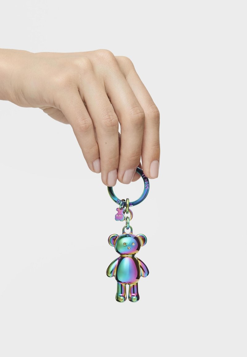 Llavero con un charm de oso de peluche iridiscente, adjunto a un anillo metálico circular, sostenido por una mano con esmalte de uñas neutro.
