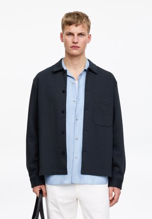 Young man wearing a black buttoned jacket over a light blue shirt, white pants, holding a black bag against a white background.