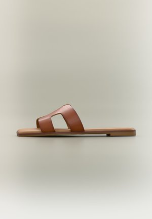 Sandale plate marron à enfiler avec une large bride en cuir présentant des découpes géométriques, sur un fond neutre uni.