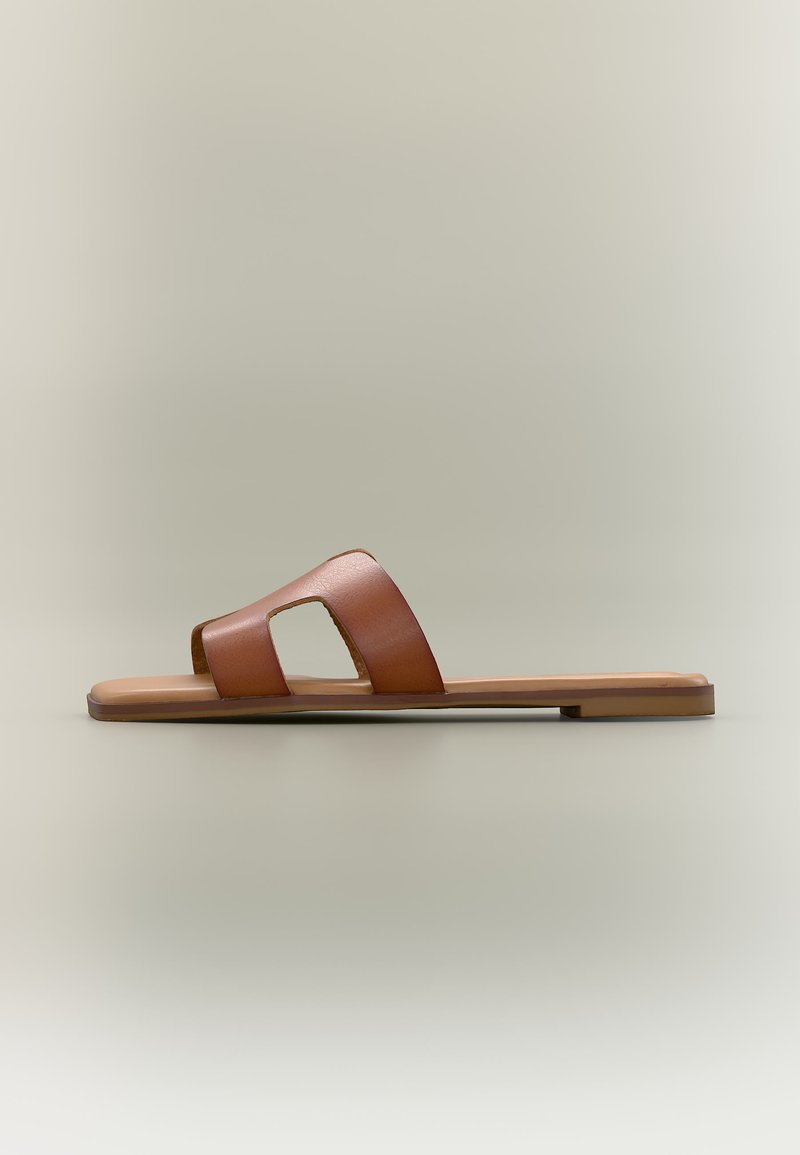 Sandale plate marron à enfiler avec une large bride en cuir présentant des découpes géométriques, sur un fond neutre uni.