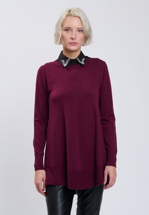 Lyshåret kvinde iført en bordeaux rullekravesweater over en sort skjorte med en udsmykket krave, kombineret med sorte læderbukser.