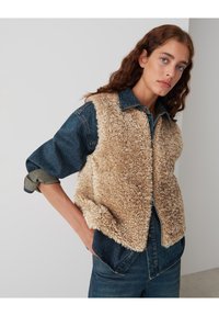 Gilet en denim avec un devant en fausse fourrure beige douce, doté d'une fermeture éclair, de manches roulées et d'un col. La texture contraste de manière saisissante.