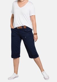 Bílé tričko s výstřihem do V ve spojení s námořnicky modrými cargo šortkami ke kolenům s ohrnutými lemy a hnědým pleteným opaskem. Celý outfit doplňují bílé tenisky.