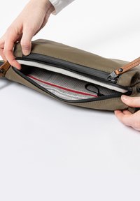 Olijfgroene clutch met een rits, heeft een lichtgrijze binnenvoering met een rasterpatroon, rode accent en een lederen strap.
