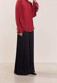 Rød gennemsigtig bluse med plissee lange ærmer og sort plissee maxi nederdel. Outfittet har en knappelukning foran og en flowy silhuet.