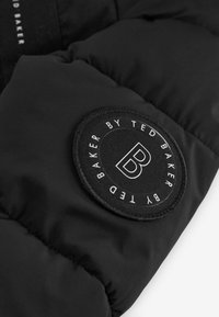 Čierna prešívaná bunda so zaoblenou čiernou látkovou nášivkou s bielym písmom a výrazným logom 'B', ktorá prezentuje hladkú textúru.