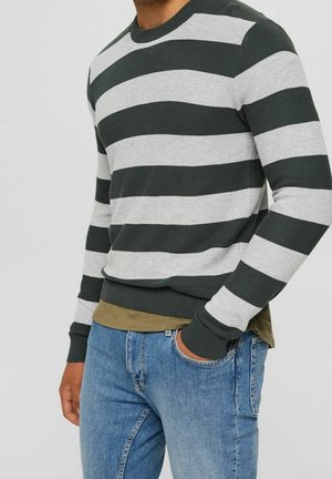 Homme portant un pull à rayures horizontales vert foncé et gris clair avec une chemise vert olive en dessous et un jean bleu.
