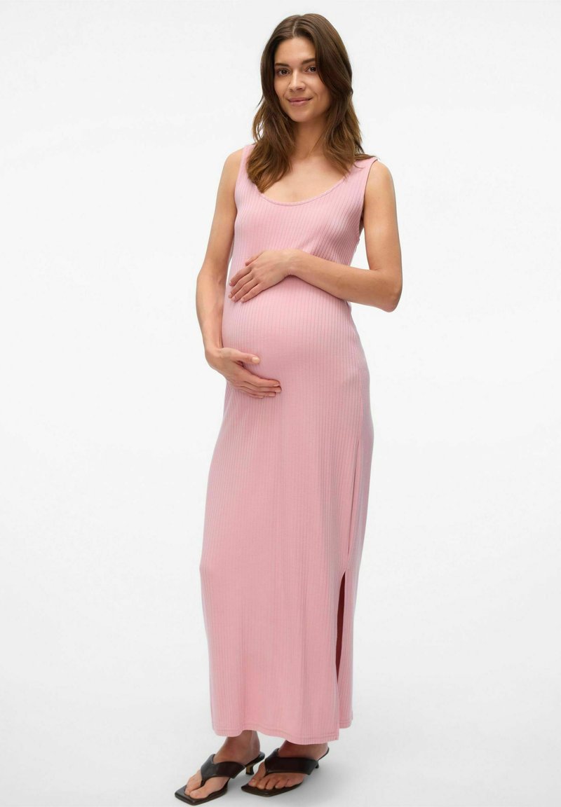 Robe de maternité rose clair côtelée avec un décolleté dégagé, fente sur le côté et une silhouette ajustée, accompagnée de tongs noires.