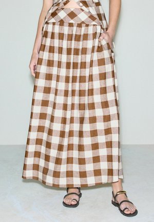 Vrouw die een bruine en witte geruite maxirok draagt, met haar hand in de zak en zwarte sandalen met open teen en een gouden enkelbandje.