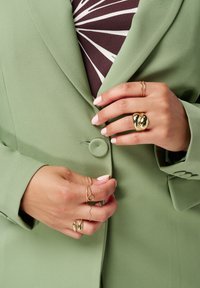 Lichtgroene op maat gemaakte blazer met een ronde knop, gedragen over een bruine top met een wit geometrisch patroon; gouden ringen en armband om de handen.