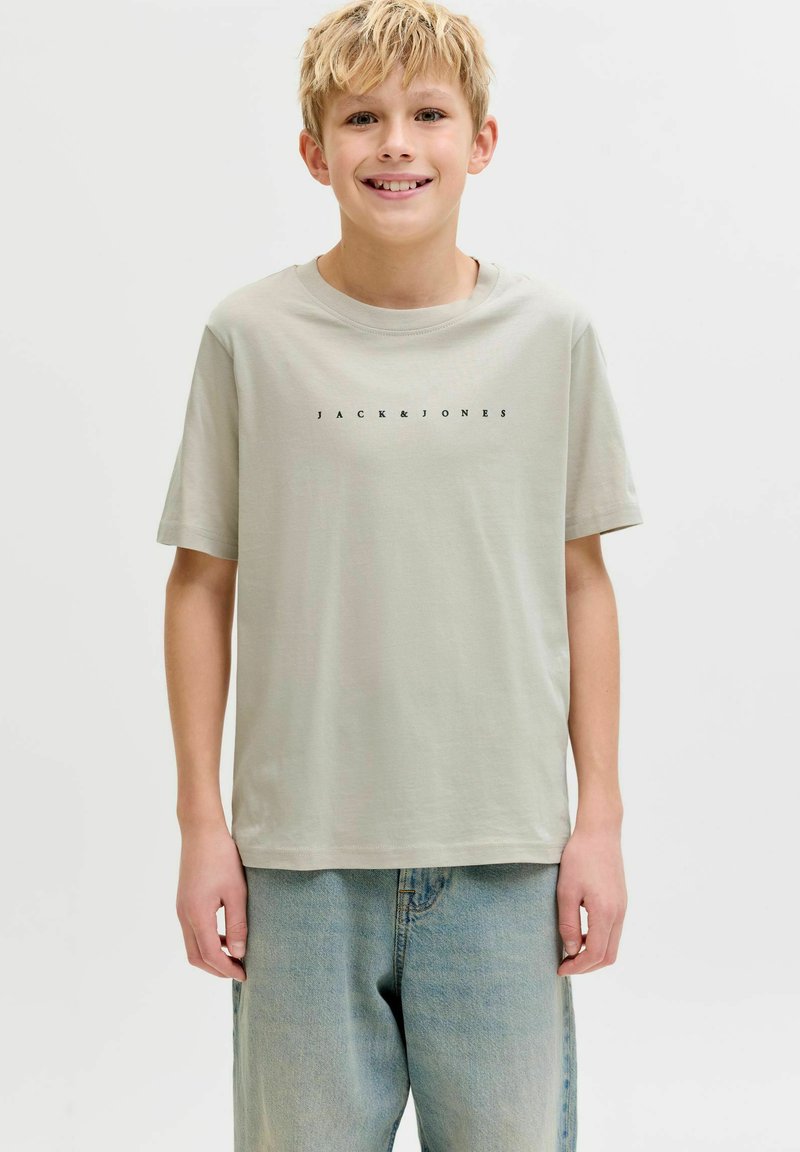 Heledate juustega poiss naeratab, kandes helerohelist Jack & Jones t-särki ja siniseid teksapükse, taustal valge tavaline sein.